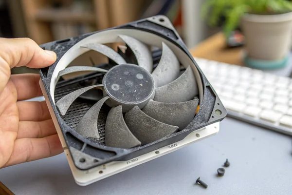A dust-clogged PC case fan removed from a computer, showing thick gray lint accumulation on the blades and around the hub, held in a hand over a desk