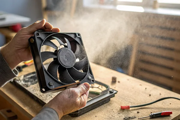 A PC case fan removed and held in a hand in a dusty industrial workshop, with visible dust particles suspended in the air near a workbench