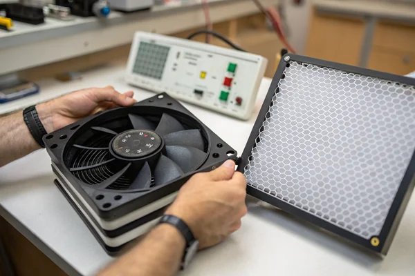 A technical engineer examining a DC axial fan on a workbench next to a mesh intake filter panel, with test equipment visible in the background