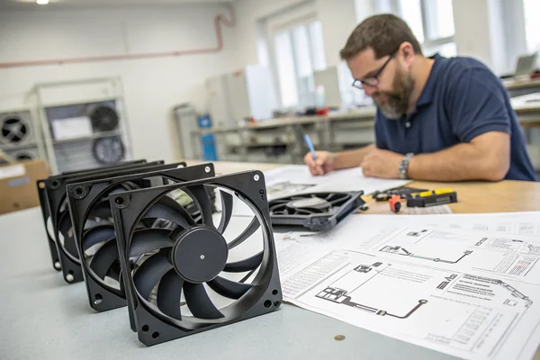 A technician at a workbench handling multiple 120mm case fans still in manufacturer packaging, comparing specifications on printed spec sheets laid flat on the surface