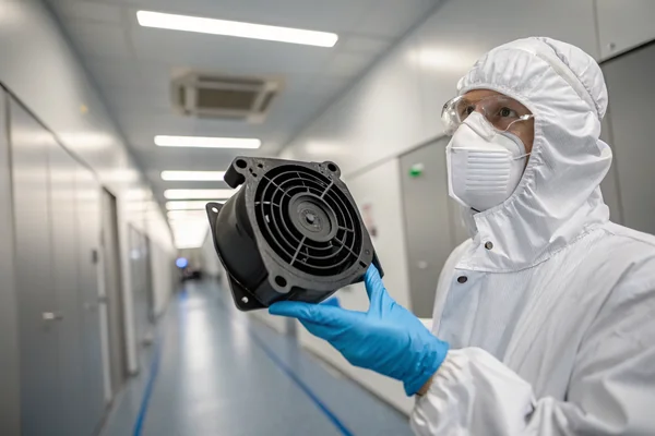 A technician in a cleanroom environment holding a small compact blower fan module with black housing and visible outlet port, inspecting it under bright overhead lighting
