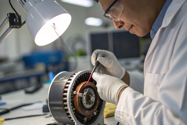 A technician in a lab environment holding a DC blower fan module with the housing removed, inspecting the bearing housing and motor windings under a workbench lamp