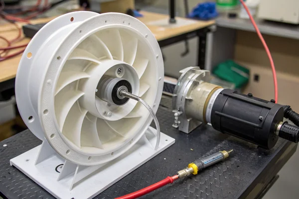 Close-up of a compact blower fan impeller spinning inside a white molded plastic volute housing, photographed during an airflow bench test with pressure tubing connected to the outlet port