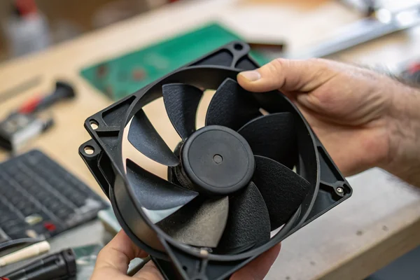 DC axial cooling fan held in a technician's hand showing blade geometry and motor hub against a workshop bench background