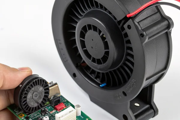 DC blower fan approximately 60mm diameter with black plastic scroll housing held in a technician's palm next to a circuit board assembly showing how it fits the compact enclosure footprint