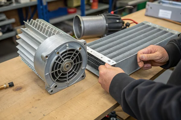 Technician at a workbench holding a compact high-pressure DC blower next to a dense aluminum heat exchanger assembly, comparing the blower inlet diameter against the exchanger's fin depth