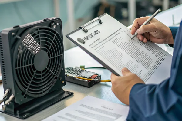 Technician reviewing printed compliance certificates and test documents next to a DC cooling fan on an inspection desk