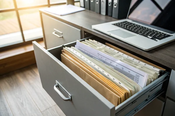 Filing cabinet drawer with organized compliance documentation folders next to a laptop on an office desk