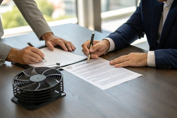 Two people signing a written supplier agreement document across a conference table with a sample fan product visible